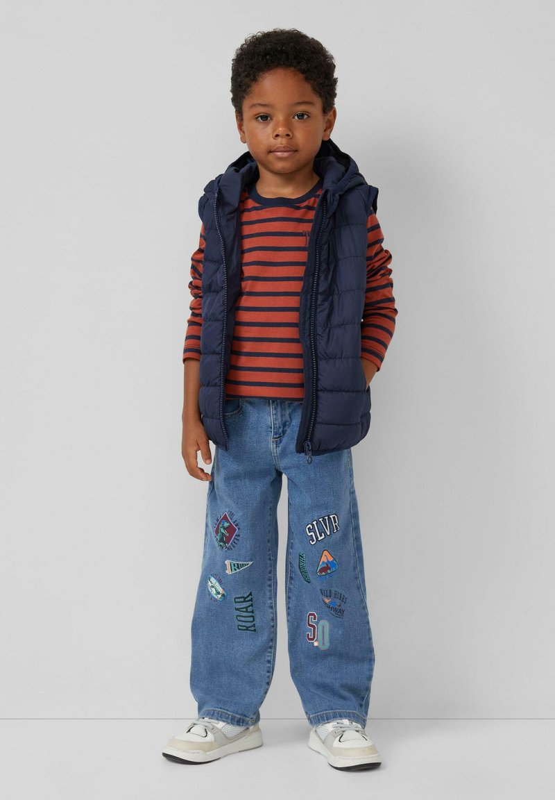 Jongentje in een marineblauwe gewatteerde vest, met een rood-oranje en marineblauwe gestreepte lange mouwen shirt, blauwe spijkerbroek met patches en witte sneakers.