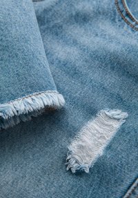 Tela de mezclilla azul claro con un dobladillo deshilachado y un parche desgastado. La textura es suave con patrones de tejido vertical y horizontal.