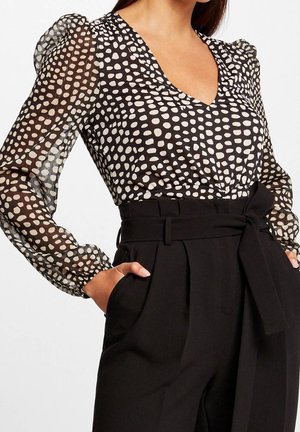Femme portant un chemisier transparent à pois noirs et blancs à manches longues, associé à un pantalon noir taille haute avec ceinture en tissu.
