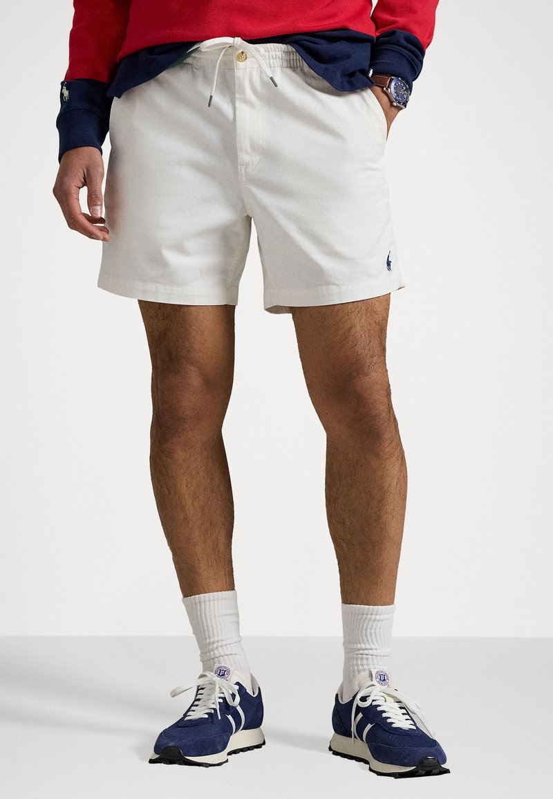 Muž v bílých šortkách na stahování, modro-červeném tričku s dlouhým rukávem, bílých žebrovaných ponožkách a modrých teniskách stojí proti jednoduchému pozadí.