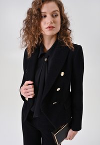 Blazer noir à chevrons avec boutons dorés, associé à un chemisier noir. Tenant une petite pochette noire. Tissu texturé et coupe ajustée.
