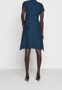 Spetsklänning i djup turkos med korta ärmar, vågig nederkant och dragkedjestängning i ryggen. Har en figurnära byst och utsvängd kjol.