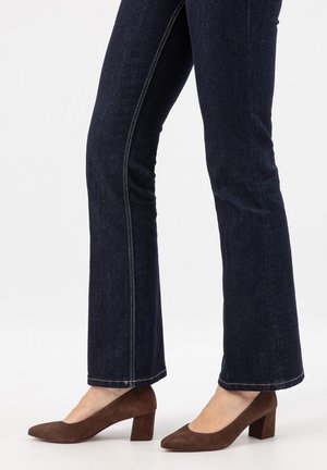 Person trägt dunkelblaue Jeans mit geradem Bein und braune Wildleder-Schuhe mit Blockabsatz, steht vor weißem Hintergrund.