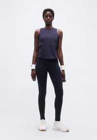 Γυναίκα με μαύρα αθλητικά ρούχα και λευκά αθλητικά παπούτσια που κρατά αλτήρες 1 κιλού, στέκεται μπροστά σε απλό ανοιχτό γκρι φόντο.