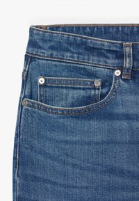 Jeans in denim di colore blu medio, design a cinque tasche, dettagli in metallo e cuciture visibili. Il tessuto presenta leggere variazioni di texture.