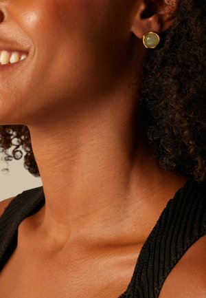 Femme aux cheveux bouclés foncés portant un haut noir texturé et une boucle d'oreille en or avec une pierre verdâtre, souriant légèrement.