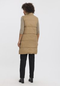 Gilet matelassé beige avec col montant, coutures horizontales et fentes latérales, associé à un pantalon noir et des bottes à talons.