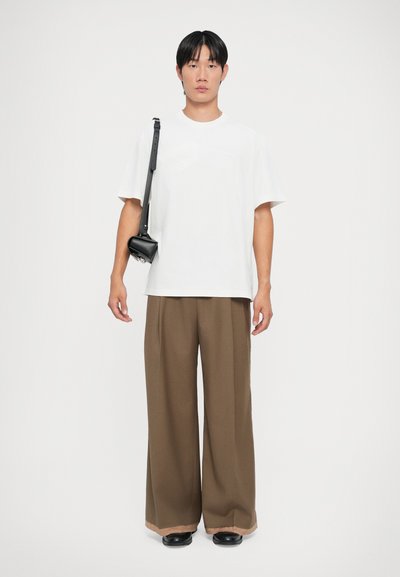 T-shirt bianco a maniche corte, pantaloni larghi marroni con un sottile dettaglio all'orlo, mentre porta una piccola borsa nera a tracolla.