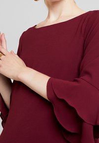 Blouse bordeaux avec un col bateau et des manches volantées. Confectionnée dans un tissu texturé, présentant une surface lisse avec un léger éclat.