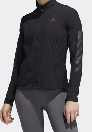 Dorosły noszący czarną sportową kurtkę na zamek błyskawiczny z logo Adidas na piersi oraz paskowane detale na lewym rękawie.