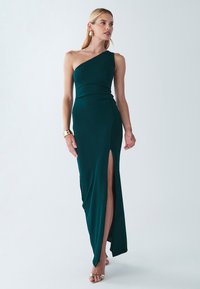 Yhden olkapään teal-mekko, jossa on sivuhires, valmistettu sileästä kankaasta. Ominaisuuksina tiukka siluetti ja elegantti laskostus. Kultainen koru korostaa.