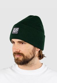 Sötétzöld kötött sapka hajtott mandzsettával, fekete téglalap alakú folttal, melyen fehér "HOMEBOY" felirat található. Puha textúrájú és bordázott kialakítású.