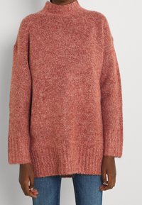 Übergroßer, strukturierten Pullover in einer sanften, rötlich-pinken Farbe. Mit hohem Kragen, herunterhängenden Schultern und geripptem Saum. Kombiniert mit blauen Jeans.