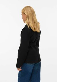 Cappotto nero con cintura, maniche lunghe, spalle strutturate e cucitura posteriore sartoriale. Abbinato a jeans blu a gamba larga.