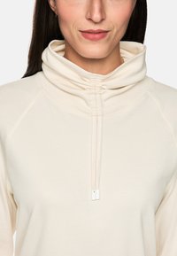 Light beige pullover with a high, gathered collar and a subtle drawstring. Made of soft, textured fabric with raglan sleeves.