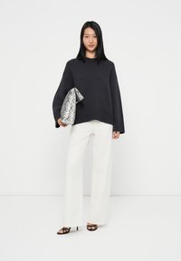 Dunkelgrauer Strickpullover mit geripptem Saum, kombiniert mit weißen, weit geschnittenen Hosen. Das Model hält eine gemusterte graue Tasche und trägt schwarze Absatzschuhe.