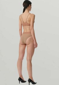 Donna in piedi di spalle, che indossa un reggiseno nudo e uno slip a vita alta con tacchi neri, su uno sfondo chiaro e semplice.