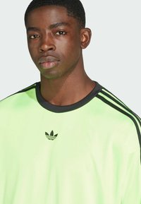 Zaļa sporta krekls ar melniem akcentiem, ar apaļu kakla izgriezumu un trim melnām svītrām gar pleciem. Mazs melns logo uz krūtīm.