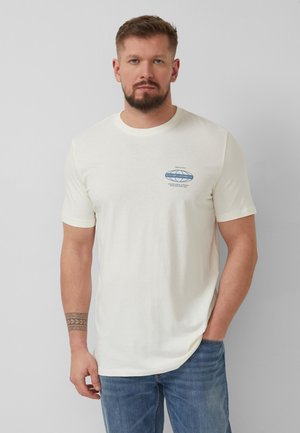 Man met baard, draagt een wit T-shirt met korte mouwen en een klein blauw logo op de borst, en een blauwe spijkerbroek, staand tegen een grijze achtergrond.