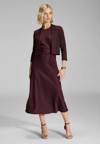 Vestido de satén burdeos con una silueta A-line hasta la rodilla, combinado con un cárdigan corto brillante y tacones negros con tiras. Detalle de torsión sutil en la cintura.