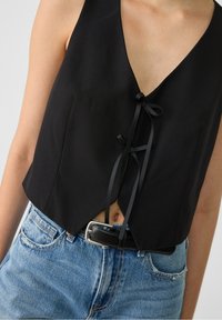 Black cropped top with a deep V-neck design, featuring a tie closure with ribbon accents. Paired with light blue high-waisted denim jeans.