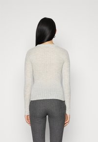 Donna con lunghi capelli neri, che indossa un maglione aderente grigio chiaro a coste e pantaloni grigio scuro a spina di pesce, di spalle su uno sfondo neutro.