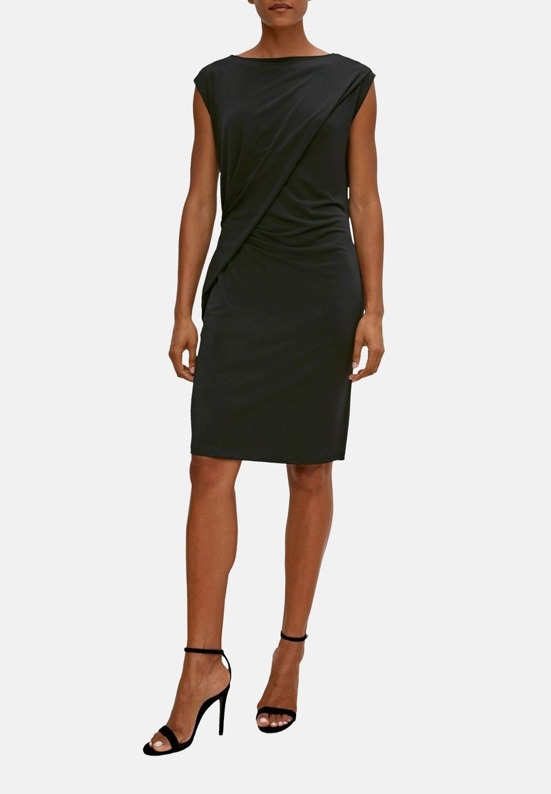 Femme portant une robe noire sans manches longueur genou avec un détail drapé et des sandales noires à talons hauts avec bride à la cheville sur un fond uni.