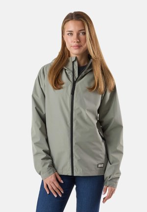 Frau mit langem hellbraunem Haar trägt eine hellgraue Jacke mit Reißverschluss und blaue Jeans, steht vor einem schlichten weißen Hintergrund.