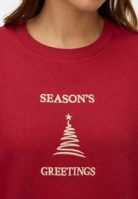 Rdeč pulover z belo izvezenim napisom "SEASON'S GREETINGS" in stiliziranim božičnim drevescem na sredini.
