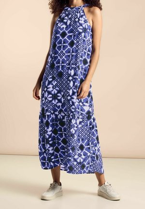 Mujer con vestido azul sin mangas de largo hasta el tobillo con estampado, combinando con zapatillas blancas, posando contra un fondo beige.