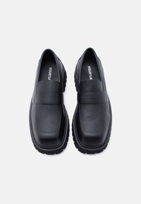 Μαύρα δερμάτινα μοκασίνια με λεία υφή, μαλακό πάτο, ανθεκτική σόλα από καουτσούκ και μινιμαλιστικό σχέδιο με οριζόντια λωρίδα κατά μήκος της πελματιαίας πλευράς.