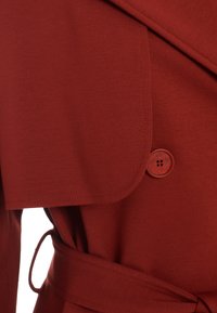 Manteau rouge avec une texture lisse, doté d'un large col, d'une fermeture à un seul bouton et d'un détail de taille nouée.