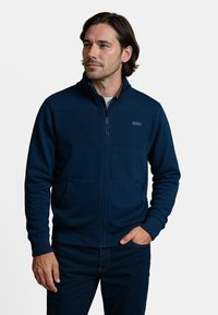 Marineblauwe rits-trui met ribbeltextuur en opstaande kraag, voorzien van twee voorzakken en een klein logo op de borst.