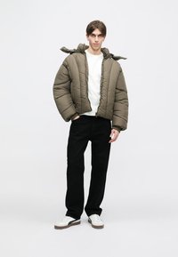 Casaco puffer caqui com colarinho alto e capuz, apresentando um design acolchoado. Usado com uma camisa branca e calças pretas, complementado com ténis.