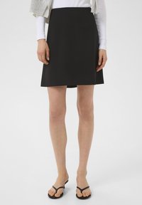 Mini-jupe noire avec une texture lisse, taille ajustée, forme évasée et longueur mi-cuisse, associée à des sandales à plat noires.
