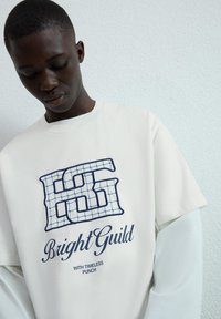 Jasnozólty bawełniany t-shirt z krótkim rękawem, ozdobiony niebieskim haftowanym logo "HG" oraz napisem "Bright Guild" w kontrastowej czcionce.
