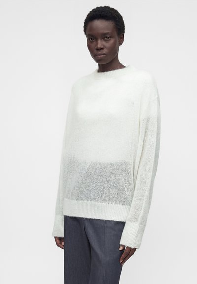 Γυναίκα με κοντά σγουρά μαλλιά που φοράει έναν άνετο λευκό χνουδωτό πουλόβερ και σκούρο γκρίζο παντελόνι, στέκεται μπροστά σε απλό φωτεινό φόντο.