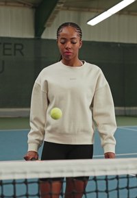 Heledad beež sweatshirth, ümmarguse kaelusega, lõdva lõikega ja logoga, kombinatsioonis mustade lühikestega. Tennisepall hõljuv võrgu kohal.