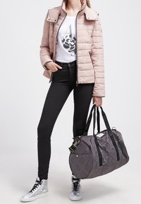 Kvinna iklädd ljusrosa vadderad jacka, vit t-shirt med grafik, svarta skinny jeans, silverfärgade sneakers, håller en stor gråmönstrad duffelväska.