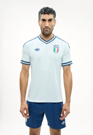 Mand iført hvid italiensk fodboldtrøje med blå kanter og matchende blå shorts, stående mod en ensfarvet hvid baggrund.