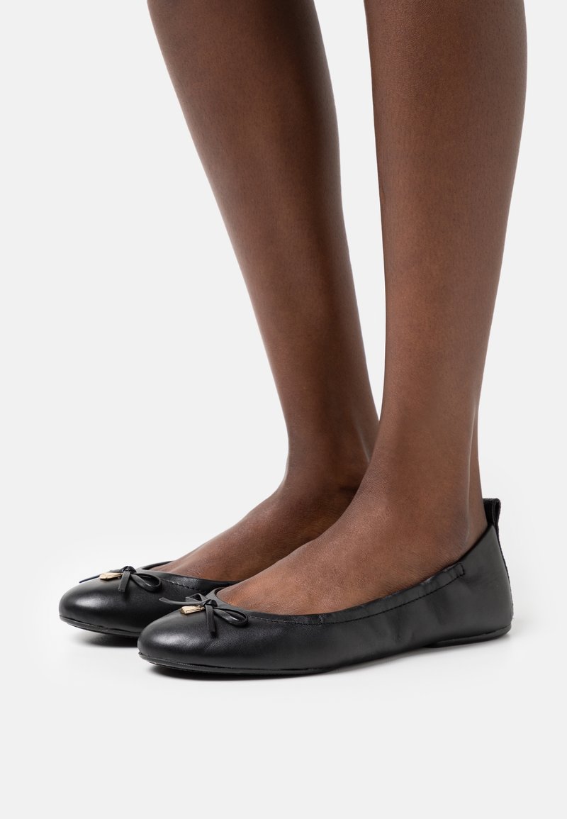 Pieds portant des ballerines en cuir noir avec de petits nœuds et des accents dorés sur un fond blanc uni.