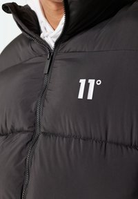 Chaqueta de plumas negra con cierre de cremallera, que presenta un logo impreso en blanco "11°" en el pecho y un cuello alto para mayor calidez.