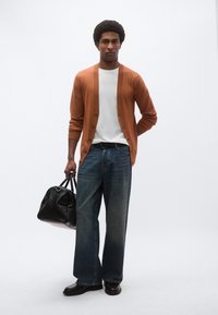 Πορτοκαλί πλεκτή ζακέτα, λευκό μπλουζάκι, μπλε τζιν παντελόνι με φαρδιά μπατζάκια και μαύρα δερμάτινα παπούτσια. Κρατά μια μαύρη υφασμάτινη τσάντα duffel. Μινιμαλιστικό φόντο.