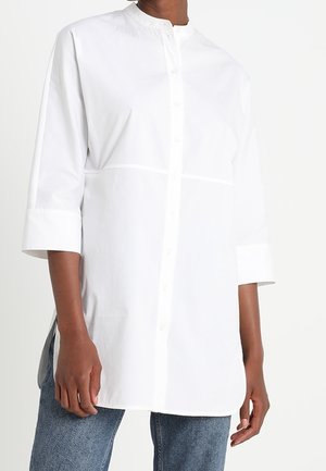 Personne portant une longue chemise blanche boutonnée avec des manches trois-quarts et un jean bleu sur un fond blanc uni.