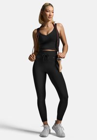 Černý sportovní crop top a legíny vyrobené z materiálu odvádějícího vlhkost, s páskem na stahování v pase a v kombinaci s bílými sportovními botami.
