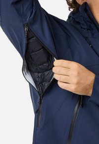 Giacca blu navy con cerniere e fodera interna trapuntata. Una mano si infila nella tasca con cerniera sul lato sinistro.