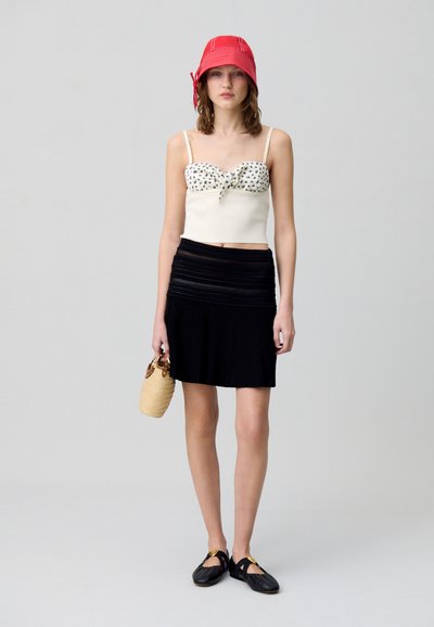 Top corto bianco con motivo floreale nero, abbinato a una gonna plissettata nera. La modella tiene in mano una piccola borsa intrecciata e indossa un cappello a secchiello rosso.
