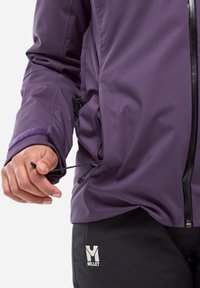 Hand adjusting side drawstring on purple jacket, wearing black pants with white Millet logo on thigh.