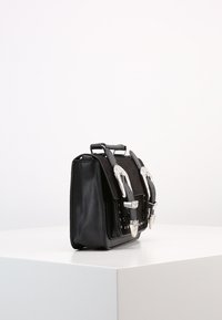 Borsa in pelle nera con due grandi fibbie argentate e un manico superiore, posizionata su un piedistallo bianco su uno sfondo bianco semplice.