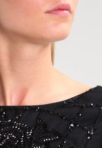 Svart topp med intrikata pärl- och paljettdetaljer, med rund halsringning och textilstruktur som framhäver axel- och halsområdet.